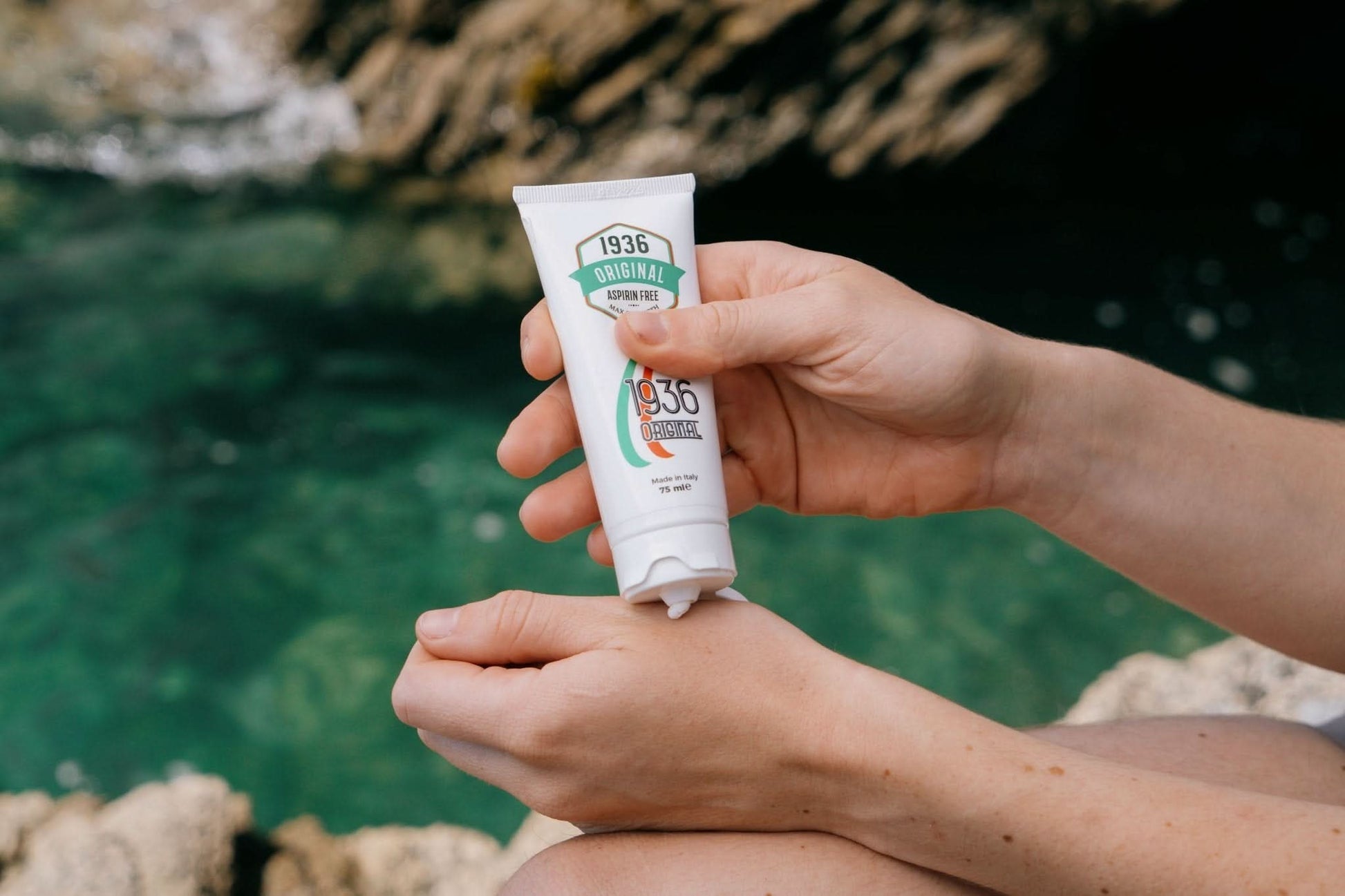 A person is applying a pain relief cream with a natural formula from a tube labeled 1936 Original onto their hand. They are sitting near a body of clear, turquoise water with rocky surroundings.